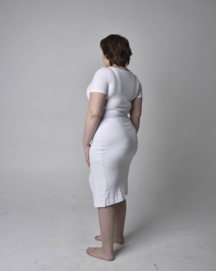 Uzun boylu, esmer kısa saçlı, dar beyaz bir vücut elbisesi giyen, hafif stüdyo arka planda ayakta duran genç bir kadının portresi..