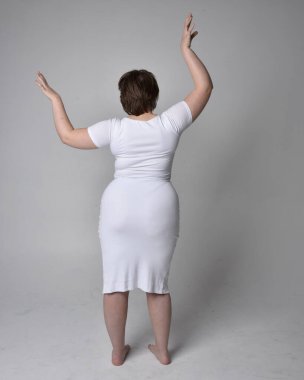 Uzun boylu, esmer kısa saçlı, dar beyaz bir vücut elbisesi giyen, hafif stüdyo arka planda ayakta duran genç bir kadının portresi..