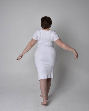 Uzun boylu, esmer kısa saçlı, dar beyaz bir vücut elbisesi giyen, hafif stüdyo arka planda ayakta duran genç bir kadının portresi..