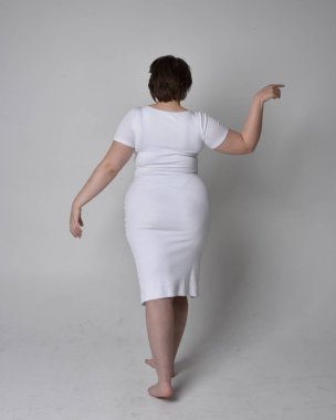 Uzun boylu, esmer kısa saçlı, dar beyaz bir vücut elbisesi giyen, hafif stüdyo arka planda ayakta duran genç bir kadının portresi..