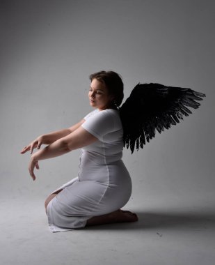 Kısa esmer saçlı, dar beyaz elbiseli ve tüylü melek kanatlarından yoksun, stüdyo arka planında gölgeli ışıklandırmayla diz çökmüş genç bir kadının tam boy portresi..