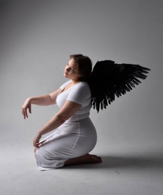 Kısa esmer saçlı, dar beyaz elbiseli ve tüylü melek kanatlarından yoksun, stüdyo arka planında gölgeli ışıklandırmayla diz çökmüş genç bir kadının tam boy portresi..