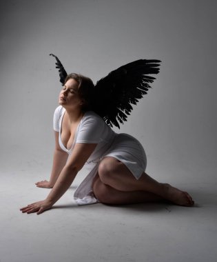 Kısa esmer saçlı, dar beyaz elbiseli ve tüylü melek kanatlarından yoksun, stüdyo arka planında gölgeli ışıklandırmayla diz çökmüş genç bir kadının tam boy portresi..