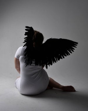 Kısa esmer saçlı, dar beyaz elbiseli ve tüylü melek kanatlarından yoksun, stüdyo arka planında gölgeli ışıklandırmayla diz çökmüş genç bir kadının tam boy portresi..