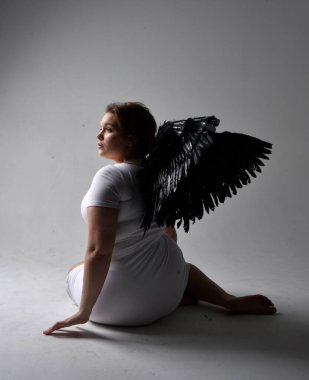 Kısa esmer saçlı, dar beyaz elbiseli ve tüylü melek kanatlarından yoksun, stüdyo arka planında gölgeli ışıklandırmayla diz çökmüş genç bir kadının tam boy portresi..