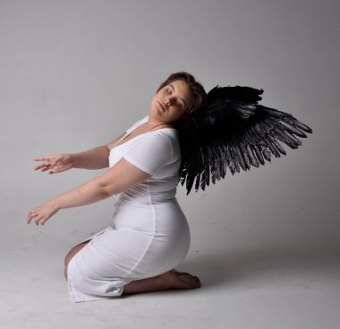 Kısa esmer saçlı, dar beyaz elbiseli ve tüylü melek kanatlarından yoksun, stüdyo arka planında diz çökmüş genç bir kadının tam boy portresi..