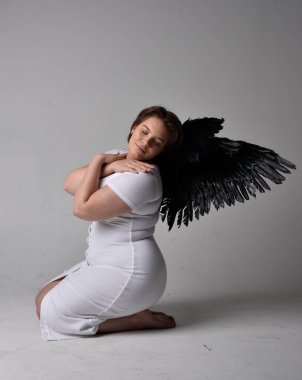 Kısa esmer saçlı, dar beyaz elbiseli ve tüylü melek kanatlarından yoksun, stüdyo arka planında diz çökmüş genç bir kadının tam boy portresi..