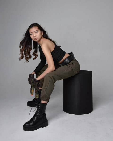Полнометражный портрет симпатичной брюнетки, азиатки в черном топе и хаки утилитарных армейских штанах и кожаных сапогах. Сидя в позе, держа в руках научно-фантастический пистолет, изолированный агент светло-серый фон студии.