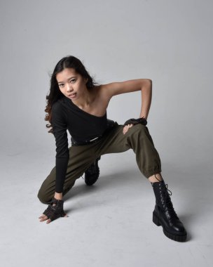 Full length portrait of pretty brunette, asian girl wearing black top and khaki utilitarian army pants and leather boots. Sitting pose , isolated agent a light grey studio background. clipart