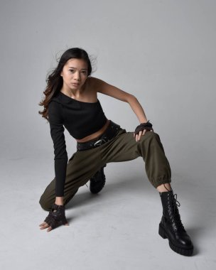 Full length portrait of pretty brunette, asian girl wearing black top and khaki utilitarian army pants and leather boots. Sitting pose , isolated agent a light grey studio background. clipart