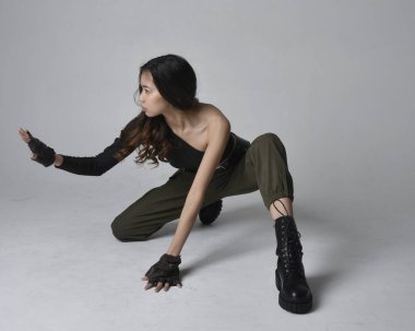 Uzun boylu, esmer, siyah bluz giyen Asyalı bir kız ve asker pantolonu ve deri çizmeleri. Oturma pozisyonu, izole bir ajan açık gri bir stüdyo arka planı..