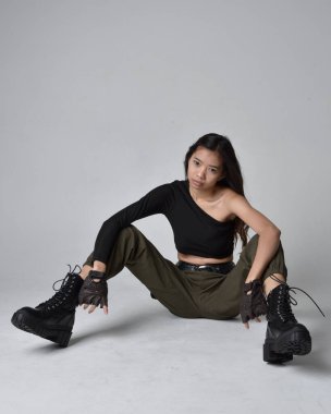 Uzun boylu, esmer, siyah bluz giyen Asyalı bir kız ve asker pantolonu ve deri çizmeleri. Oturma pozisyonu, izole bir ajan açık gri bir stüdyo arka planı..