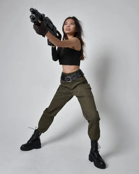 全长肖像漂亮的黑发 亚洲女孩穿着黑色上衣和卡其布军裤和皮靴 举着科幻小说枪 孤立无援的特工 背景浅灰色 — 图库照片