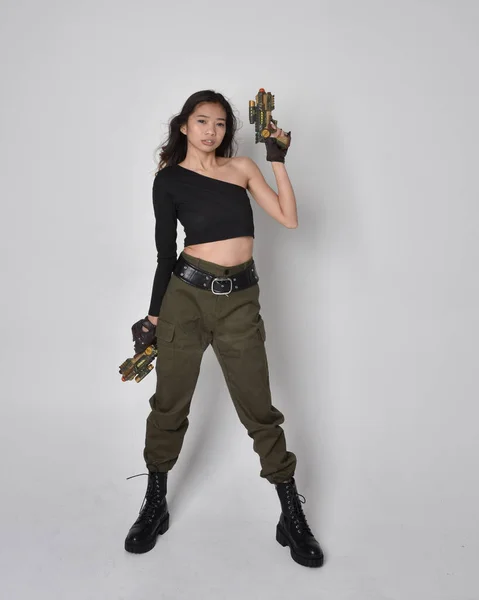 かなりブルネットの完全な長さの肖像画 黒の上とカーキ実用的な軍のズボンと革のブーツを身に着けているアジアの女の子 Sf銃を持っている立ちポーズ 孤立したエージェントライトグレーのスタジオの背景 — ストック写真