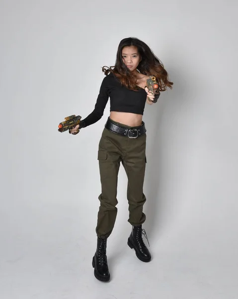かなりブルネットの完全な長さの肖像画 黒の上とカーキ実用的な軍のズボンと革のブーツを身に着けているアジアの女の子 Sf銃を持っている立ちポーズ 孤立したエージェントライトグレーのスタジオの背景 — ストック写真