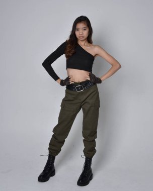 Uzun boylu, esmer, siyah bluz giyen Asyalı bir kız ve asker pantolonu ve deri çizmeleri. Hareketli ellerle poz vermek, kameradan uzaklaşmak. Açık gri bir arka planda izole edilmiş..