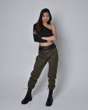 Uzun boylu, esmer, siyah bluz giyen Asyalı bir kız ve asker pantolonu ve deri çizmeleri. Hareketli ellerle poz vermek, kameradan uzaklaşmak. Açık gri bir arka planda izole edilmiş..