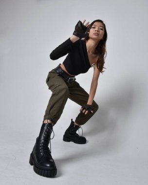 Full length portrait of pretty brunette, asian girl wearing black top and khaki utilitarian army pants and leather boots. Standing pose with gestural hands, isolated against a light grey studio background. clipart