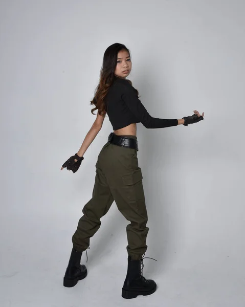 stock image Full length portrait of pretty brunette, asian girl wearing black top and khaki utilitarian army pants and leather boots. Standing pose with gestural hands, walking away from the camera.  isolated against a light grey studio background.