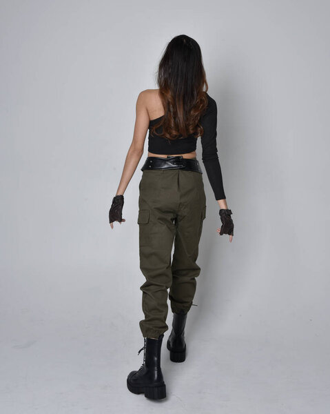 Полнометражный портрет симпатичной брюнетки, азиатки в черном топе и хаки утилитарных армейских штанах и кожаных сапогах. Стоя в позе жестом, уходя от камеры. изолированный на светло-сером студийном фоне.