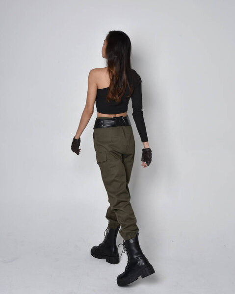 Полнометражный портрет симпатичной брюнетки, азиатки в черном топе и хаки утилитарных армейских штанах и кожаных сапогах. Стоя в позе жестом, уходя от камеры. изолированный на светло-сером студийном фоне.