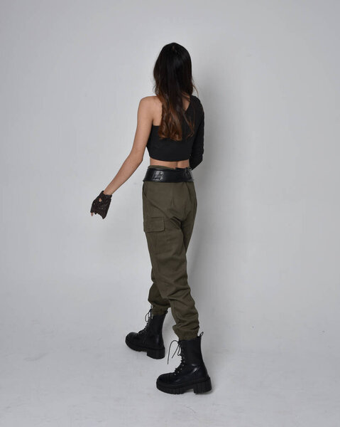 Полнометражный портрет симпатичной брюнетки, азиатки в черном топе и хаки утилитарных армейских штанах и кожаных сапогах. Стоя в позе жестом, уходя от камеры. изолированный на светло-сером студийном фоне.