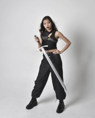 Full length portrait of pretty young asian girl wearing black tank top, utilitarian  pants and leather boots. Standing pose  holding a sword, isolated against a  studio background.