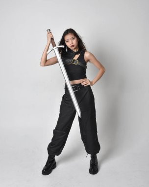 Full length portrait of pretty young asian girl wearing black tank top, utilitarian  pants and leather boots. Standing pose  holding a sword, isolated against a  studio background.