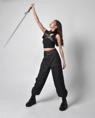 Full length portrait of pretty young asian girl wearing black tank top, utilitarian  pants and leather boots. Standing pose  holding a sword, isolated against a  studio background.