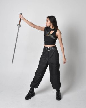 Full length portrait of pretty young asian girl wearing black tank top, utilitarian  pants and leather boots. Standing pose  holding a sword, isolated against a  studio background.
