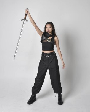 Full length portrait of pretty young asian girl wearing black tank top, utilitarian  pants and leather boots. Standing pose  holding a sword, isolated against a  studio background.