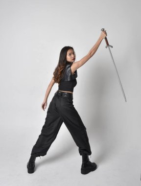Full length portrait of pretty young asian girl wearing black tank top, utilitarian  pants and leather boots. Standing pose  holding a sword, isolated against a  studio background.