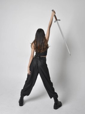 Full length portrait of pretty young asian girl wearing black tank top, utilitarian  pants and leather boots. Standing pose  holding a sword, isolated against a  studio background.