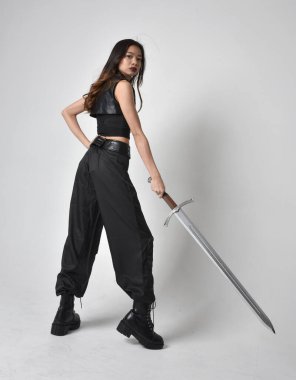 Full length portrait of pretty young asian girl wearing black tank top, utilitarian  pants and leather boots. Standing pose  holding a sword, isolated against a  studio background.