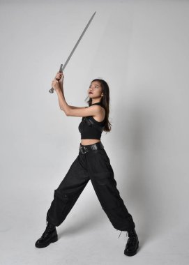 Full length portrait of pretty young asian girl wearing black tank top, utilitarian  pants and leather boots. Standing pose  holding a sword, isolated against a  studio background.
