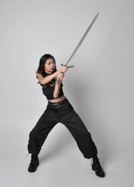 Full length portrait of pretty young asian girl wearing black tank top, utilitarian  pants and leather boots. Standing pose  holding a sword, isolated against a  studio background.