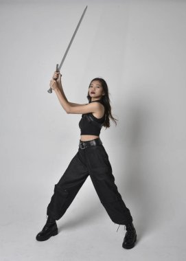 Full length portrait of pretty young asian girl wearing black tank top, utilitarian  pants and leather boots. Standing pose  holding a sword, isolated against a  studio background.