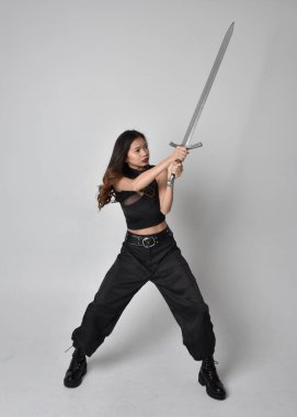 Full length portrait of pretty young asian girl wearing black tank top, utilitarian  pants and leather boots. Standing pose  holding a sword, isolated against a  studio background.