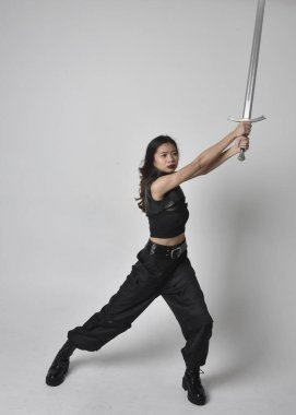 Full length portrait of pretty young asian girl wearing black tank top, utilitarian  pants and leather boots. Standing pose  holding a sword, isolated against a  studio background.