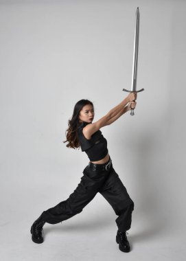 Full length portrait of pretty young asian girl wearing black tank top, utilitarian  pants and leather boots. Standing pose  holding a sword, isolated against a  studio background.