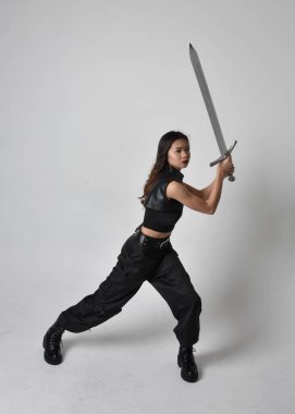 Full length portrait of pretty young asian girl wearing black tank top, utilitarian  pants and leather boots. Standing pose  holding a sword, isolated against a  studio background.
