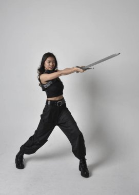 Full length portrait of pretty young asian girl wearing black tank top, utilitarian  pants and leather boots. Standing pose  holding a sword, isolated against a  studio background.