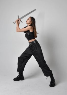Full length portrait of pretty young asian girl wearing black tank top, utilitarian  pants and leather boots. Standing pose  holding a sword, isolated against a  studio background.
