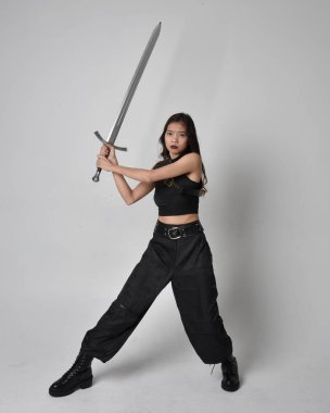 Full length portrait of pretty young asian girl wearing black tank top, utilitarian  pants and leather boots. Standing pose  holding a sword, isolated against a  studio background.