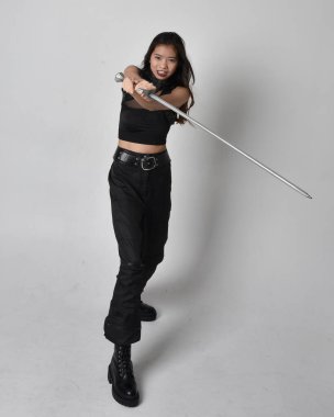 Full length portrait of pretty young asian girl wearing black tank top, utilitarian  pants and leather boots. Standing pose  holding a sword, isolated against a  studio background.