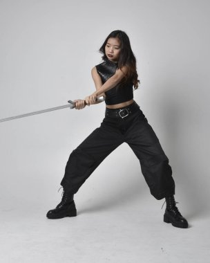 Full length portrait of pretty young asian girl wearing black tank top, utilitarian  pants and leather boots. Standing pose  holding a sword, isolated against a  studio background.