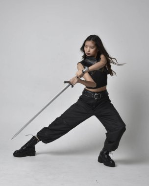 Full length portrait of pretty young asian girl wearing black tank top, utilitarian  pants and leather boots. Standing pose  holding a sword, isolated against a  studio background.