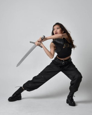 Full length portrait of pretty young asian girl wearing black tank top, utilitarian  pants and leather boots. Standing pose  holding a sword, isolated against a  studio background.