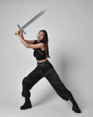 Full length portrait of pretty young asian girl wearing black tank top, utilitarian  pants and leather boots. Standing pose  holding a sword, isolated against a  studio background.