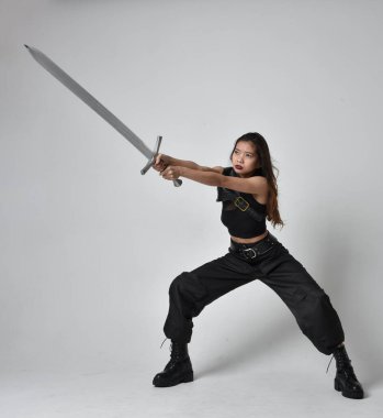Full length portrait of pretty young asian girl wearing black tank top, utilitarian  pants and leather boots. Standing pose  holding a sword, isolated against a  studio background.